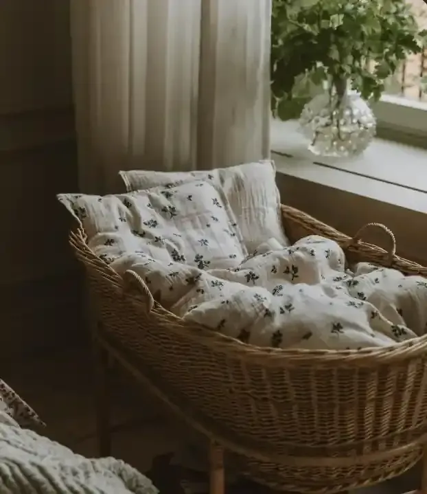 Cuna moisés de mimbre con ropa de cama floral junto a una ventana, al lado de una cortina vaporosa y una planta en maceta.