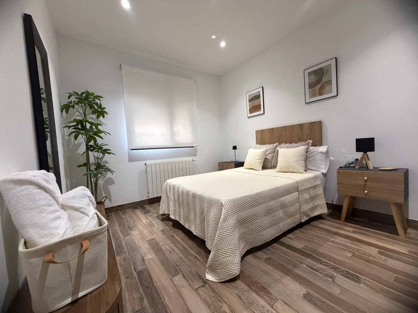 Dormitorio luminoso con cama de matrimonio, mesillas de noche de madera, silla, planta y ventana con persianas