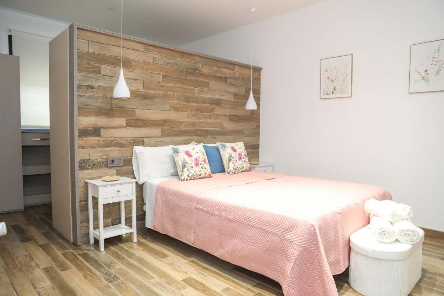 Dormitorio luminoso con una colcha rosa, una pared de madera decorativa y una mesita auxiliar debajo de un cuadro