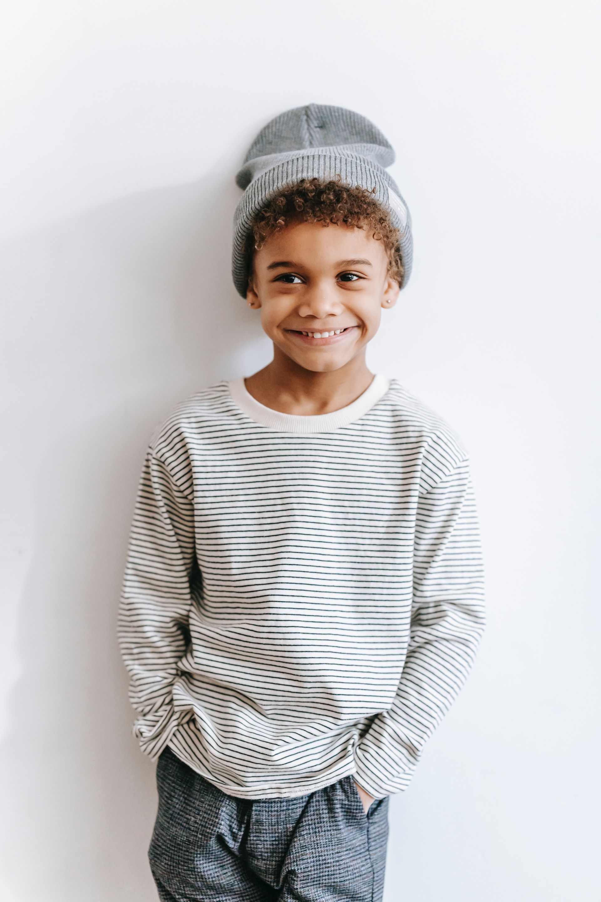 Un niño pequeño que viste una camisa de rayas y un gorro está parado frente a una pared blanca.