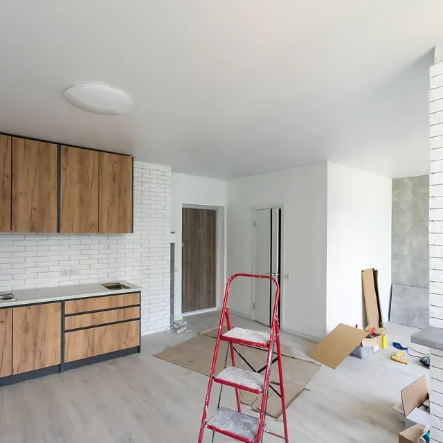 Cocina en remodelación, con gabinetes de madera, ladrillo blanco, escalera roja y materiales de construcción.