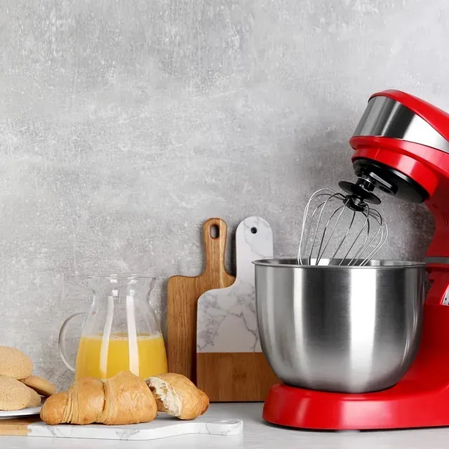 Batidora de pedestal roja con accesorio para batir, croissants, jugo y tablas de cortar sobre una encimera.