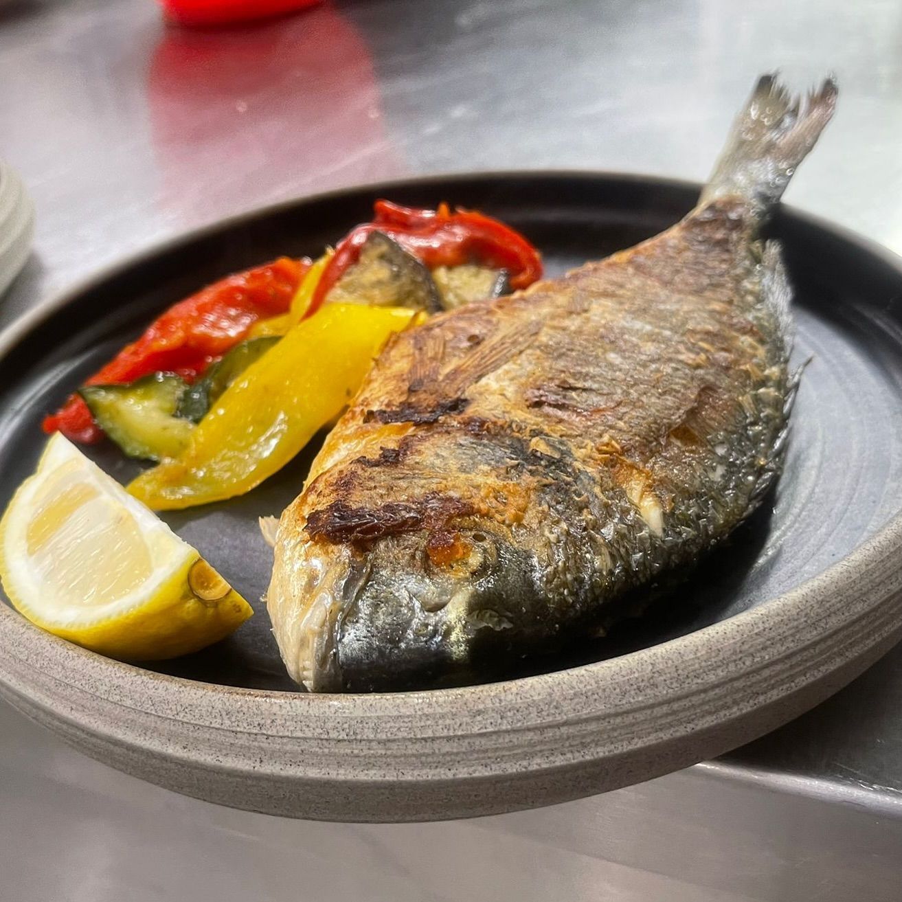 Gegrillter Fisch mit geröstetem Gemüse und Zitronenspalte auf einem dunklen Teller.