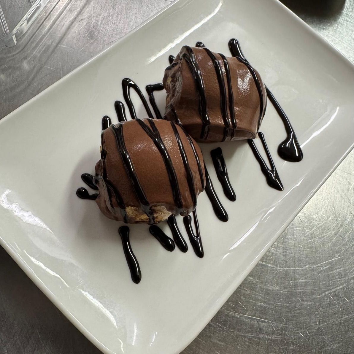 Zwei mit Schokolade überzogene Desserts, beträufelt mit Schokoladensoße, auf einem weißen rechteckigen Teller.