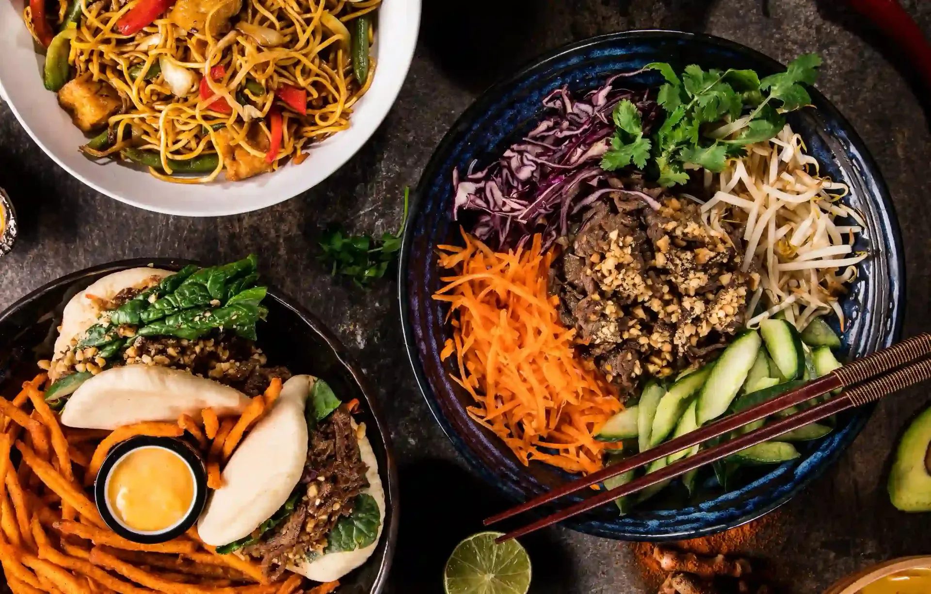 Tres cuencos de coloridos platos asiáticos de fideos y arroz con verduras y palillos sobre una mesa oscura.