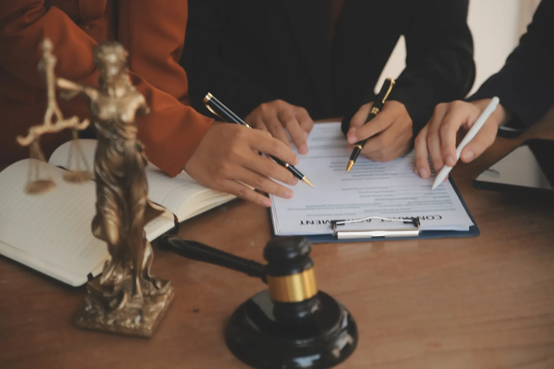 Manos firmando documentos legales con un mazo y una estatua de Lady Justice sobre un escritorio.