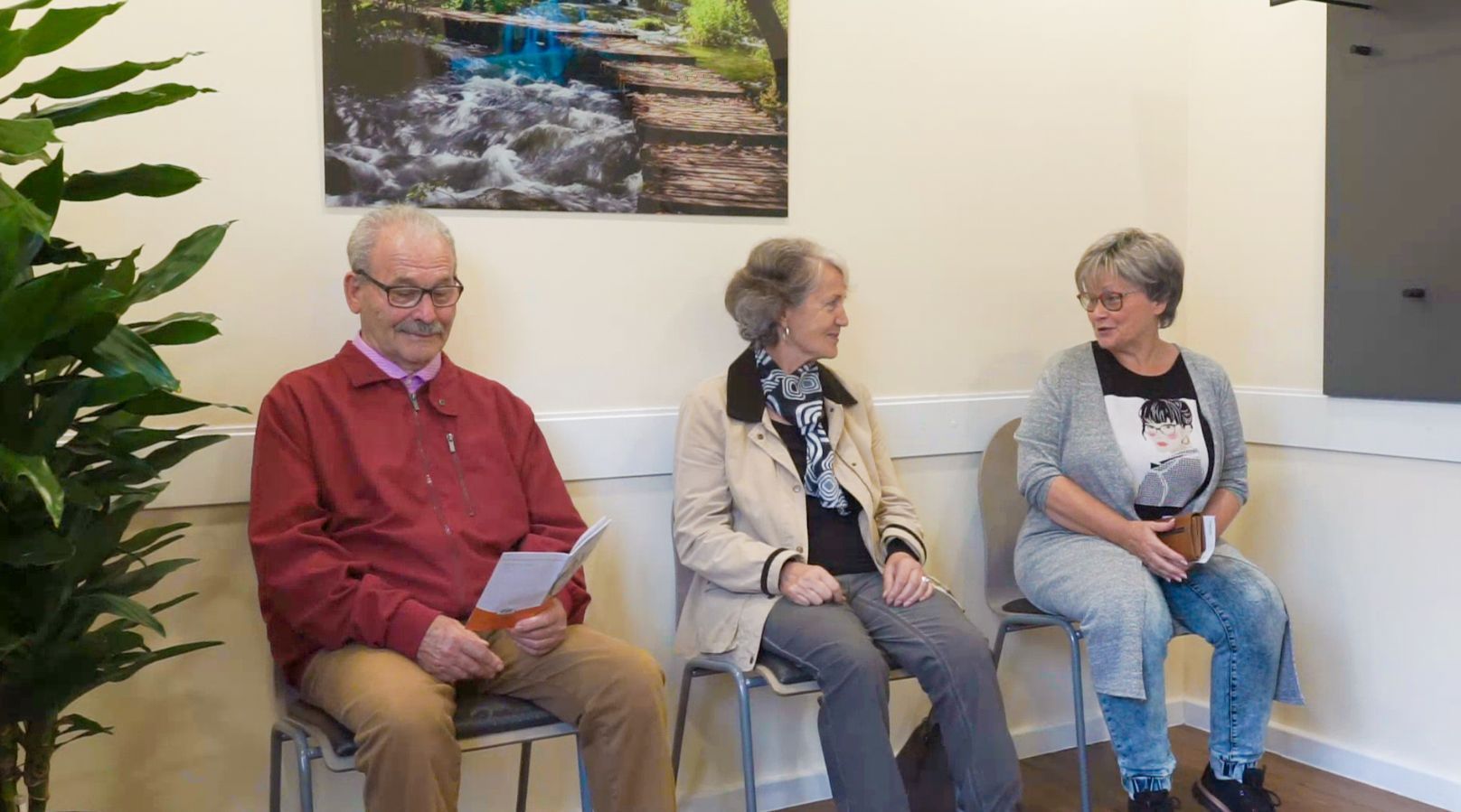 Drei Personen sitzen auf Stühlen in einem Wartezimmer.