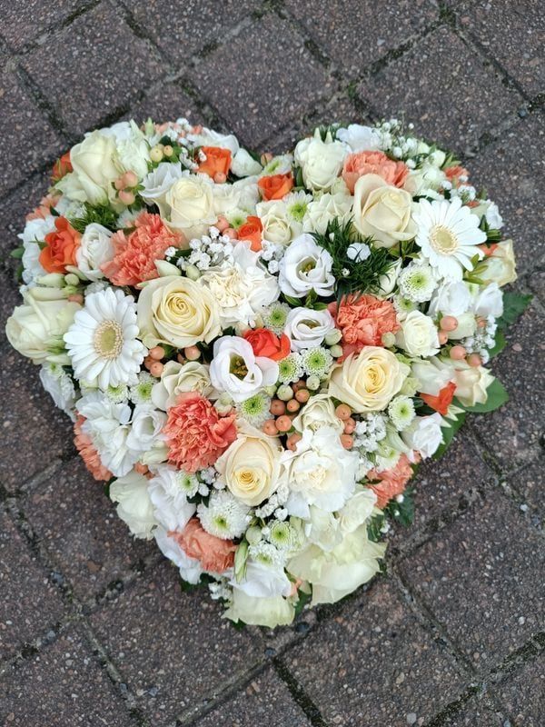 Composition florale en forme de cœur ; fleurs crème, blanc et orange sur un fond de briques.
