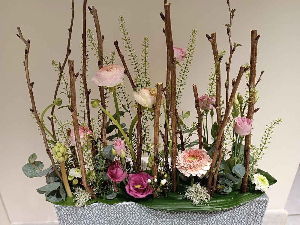 Composition florale dans un contenant gris texturé ; des fleurs rose, violet et pêche, des brindilles et de la verdure.