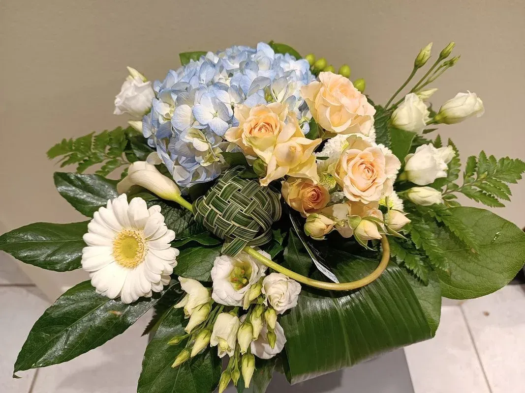 Composition florale avec des hortensias bleu clair, des roses pêche, des marguerites blanches et de la verdure.