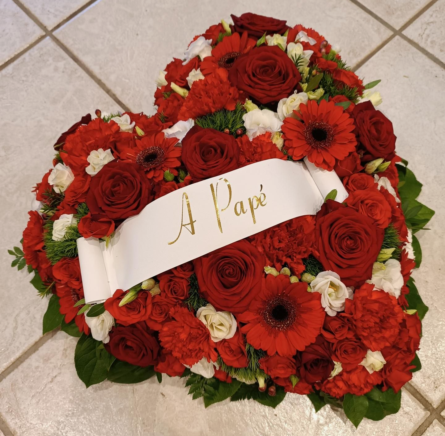 Composition florale en forme de cœur avec des roses rouges, des œillets et une banderole « A Papé », disposée sur un sol carrelé.