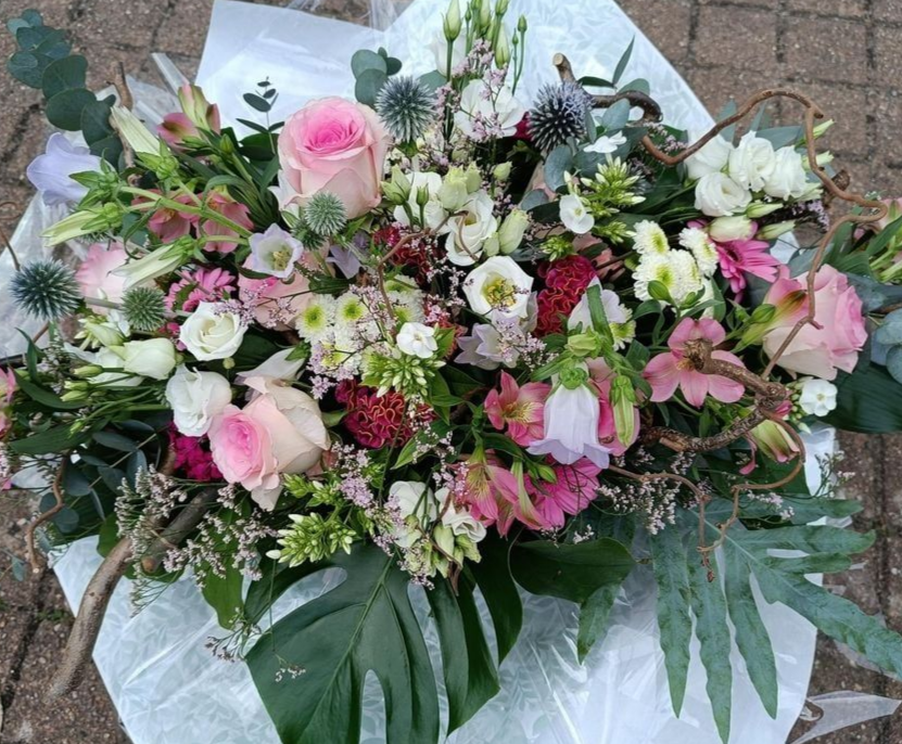 Gros plan sur un bouquet composé de roses roses, de fleurs blanc et violet et de feuilles vertes, le tout enveloppé dans du papier blanc.