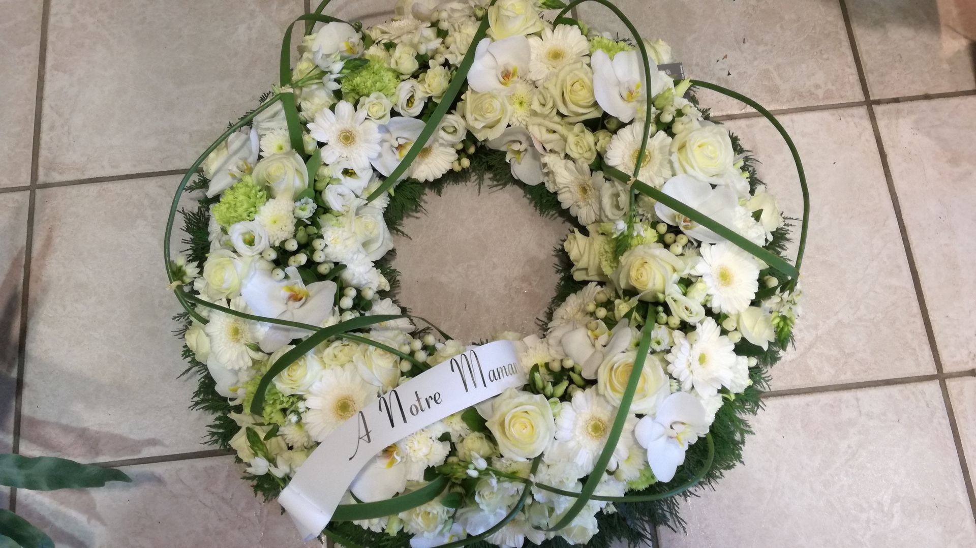 Couronne de fleurs blanches avec une banderole portant l'inscription « À votre mère » sur une surface carrelée.