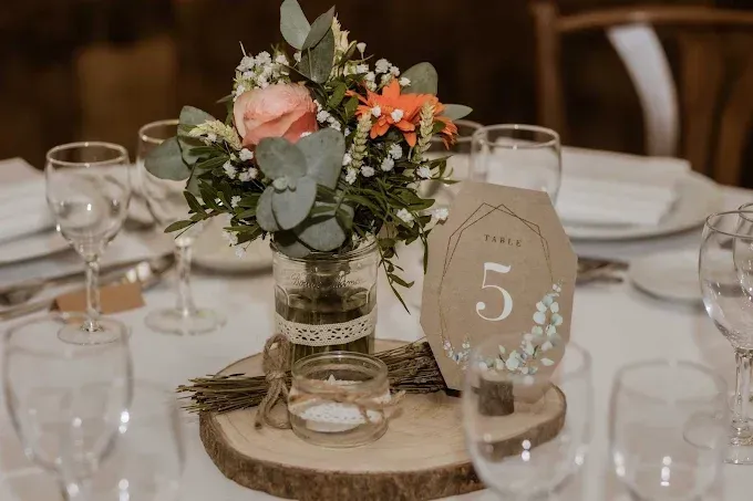 Décoration de table pour un mariage : centre de table floral, numéro de table 5, verres à vin, tranche de bois rustique.