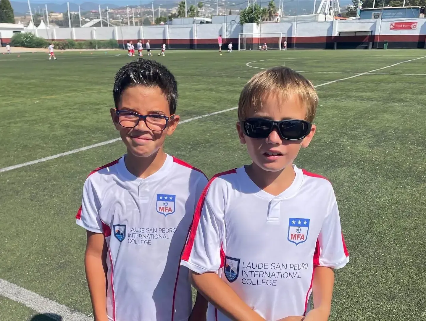 Dos jóvenes que llevan gafas de sol están parados uno al lado del otro en un campo de fútbol.