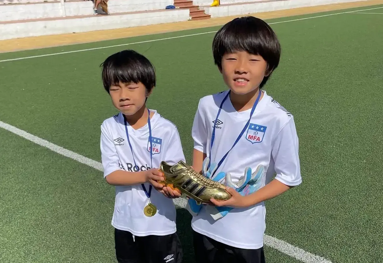 Dos niños pequeños están parados en un campo de fútbol sosteniendo botines de fútbol.