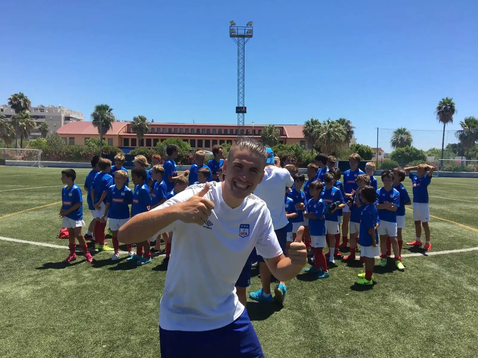 Un hombre hace un gesto de aprobación con el pulgar delante de un grupo de niños en un campo de fútbol.