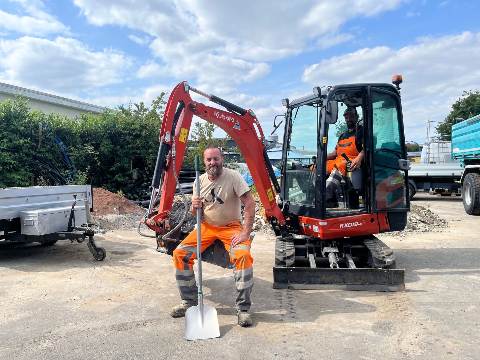 Ein Arbeiter in orangefarbener Schutzkleidung steht neben einem roten Minibagger auf einer Baustelle.