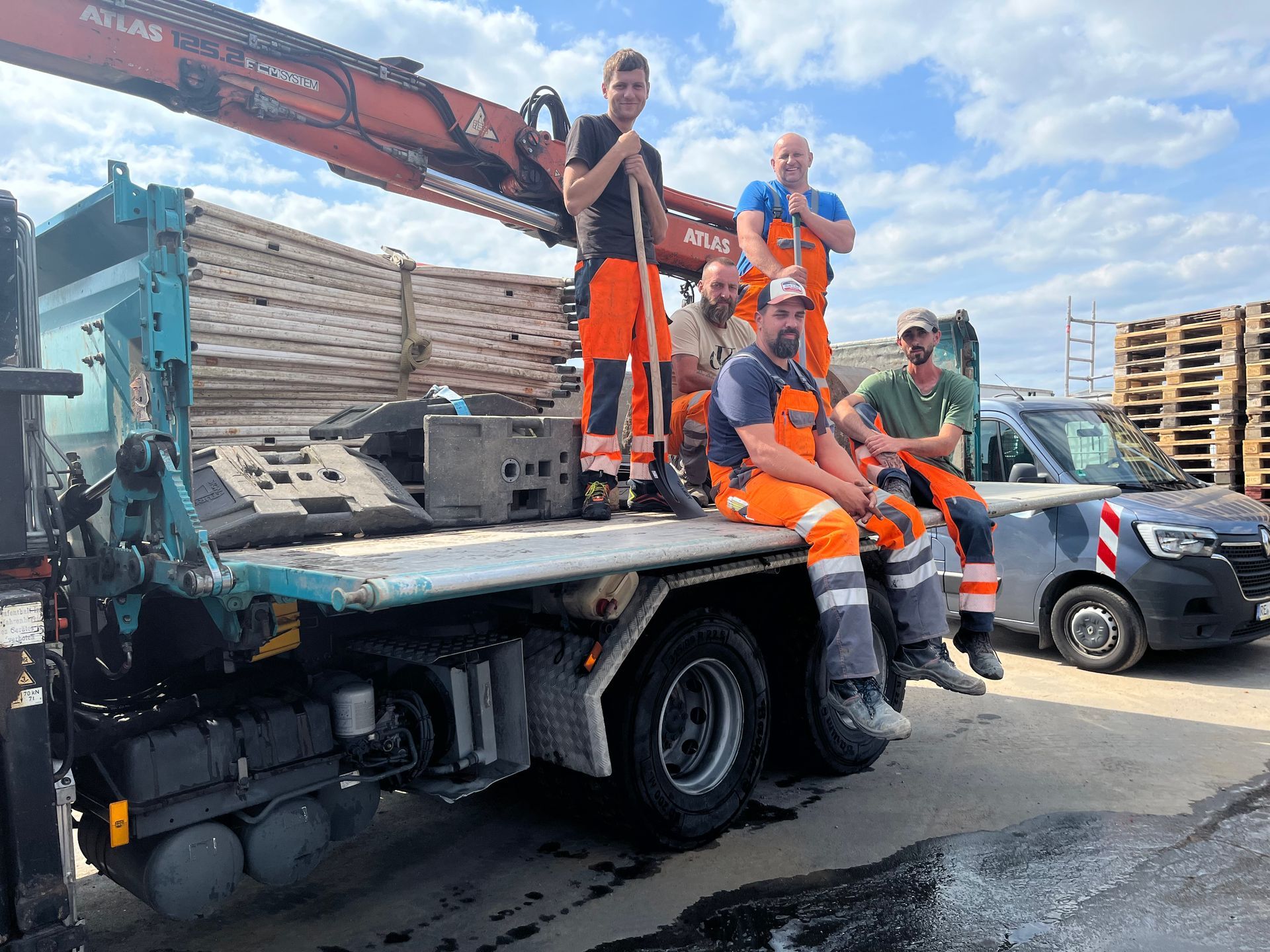 Arbeiter in orangefarbenen Arbeitskleidung auf einem Pritschenwagen auf einer Baustelle, in deren Nähe sich ein Kran und gestapeltes Baumaterial befinden.