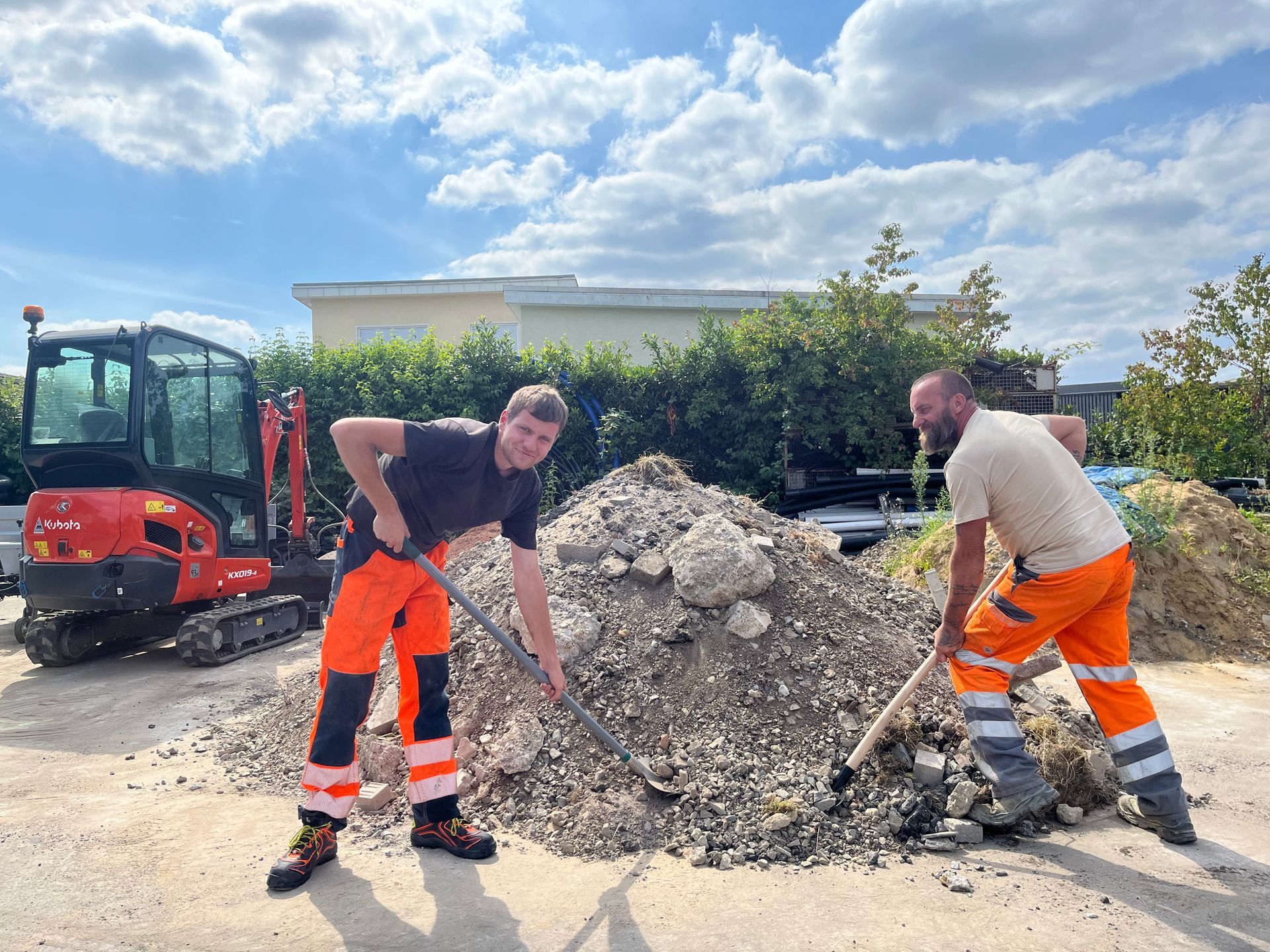 Zwei Arbeiter in orangefarbenen Hosen schaufeln Kies neben einem roten Bagger auf einer Baustelle.