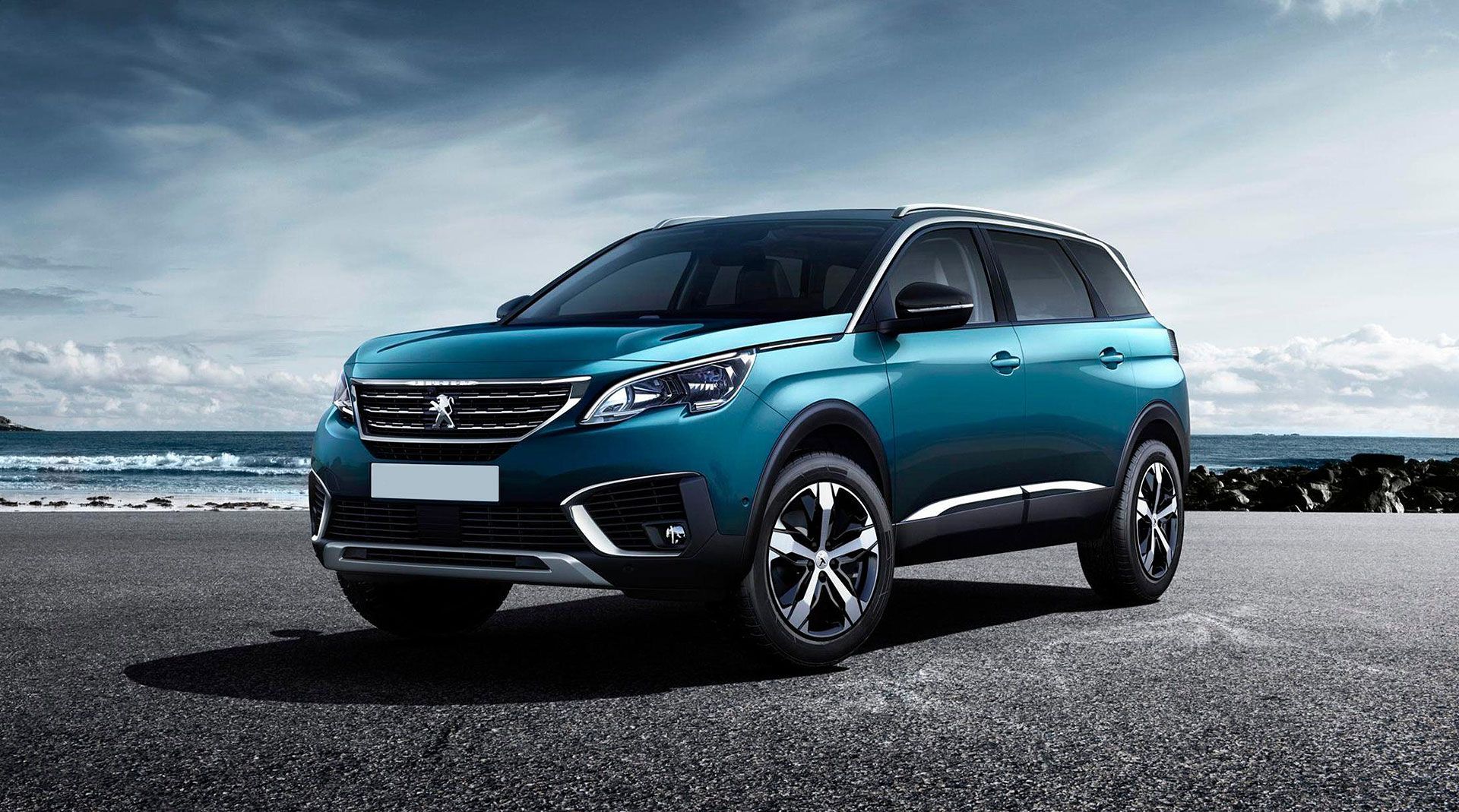 Un SUV Peugeot 5008 turquoise est garé sur une route goudronnée en bord de mer, sous un ciel nuageux.