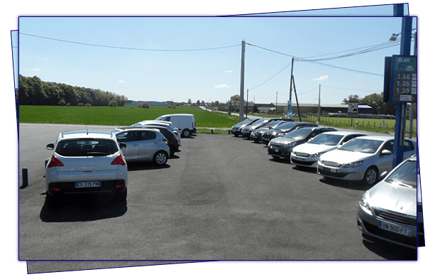 Des voitures garées sur un parking sous un ciel bleu, près d'un champ et d'arbres.