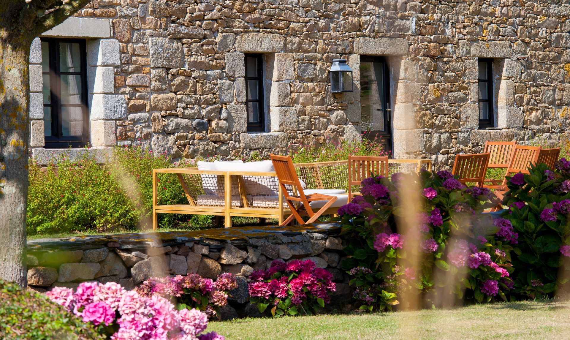Maison en pierre avec un muret et des fleurs roses plantées devant Cour intérieure d'un bâtiment en pierre avec banc en pierre, chaises et un jardin fleuri coloré à l'avant.