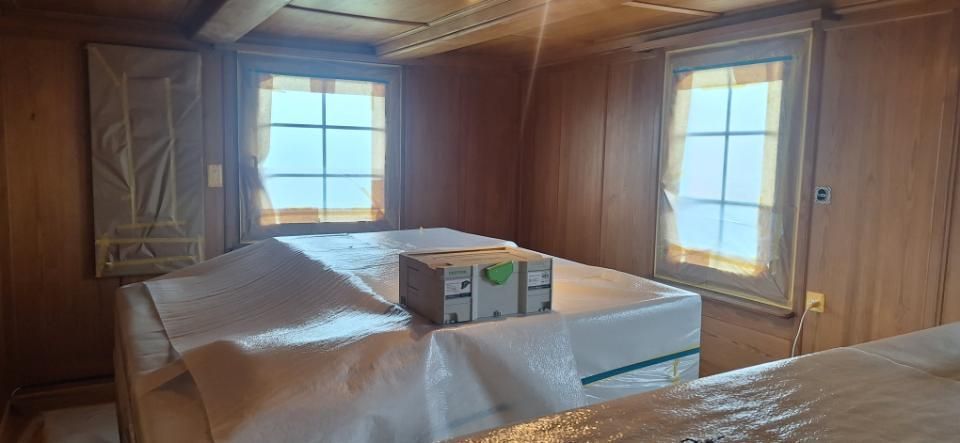 Holzgetäfeltes Zimmer mit zwei Betten an den Fenstern, eines der Betten mit Plastikfolie abgedeckt und mit einer Kiste darauf.