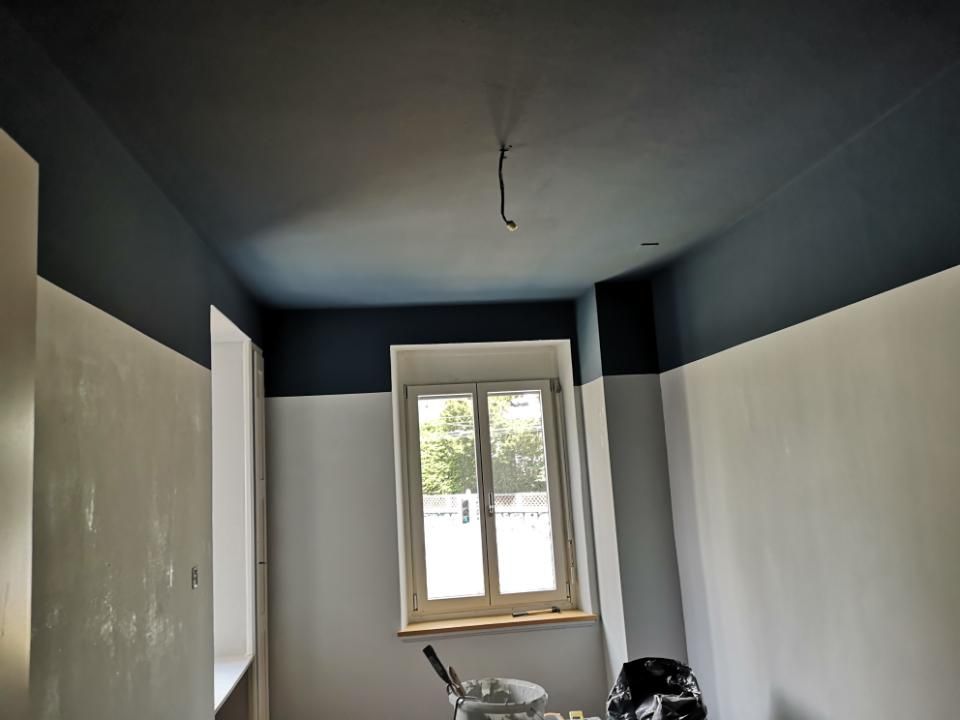 Schmaler Raum mit dunkelblauen oberen Wänden, weissen unteren Wänden, einem Fenster und freiliegenden Lichtleitungen an der Decke.