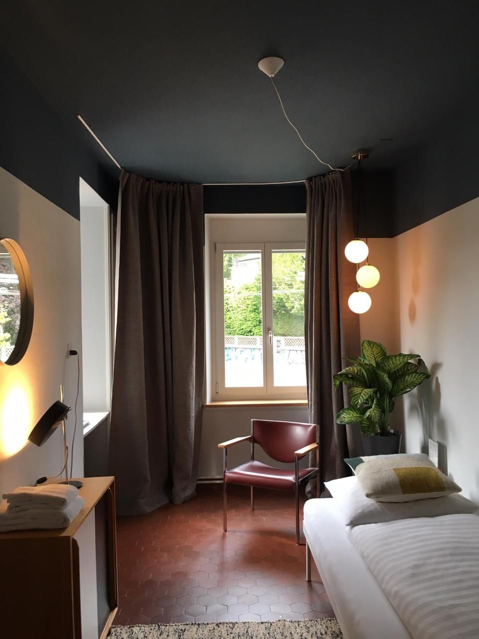 Gemütliches, modernes Schlafzimmer mit dunklen Wänden, verhängtem Fenster, weissem Bett, Stuhl und Topfpflanze