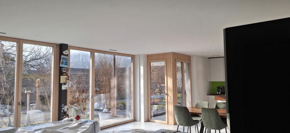 Helles, modernes Wohnzimmer mit bodentiefen Fenstern, Balkontüren und einem Esstisch an einer dunklen Akzentwand.