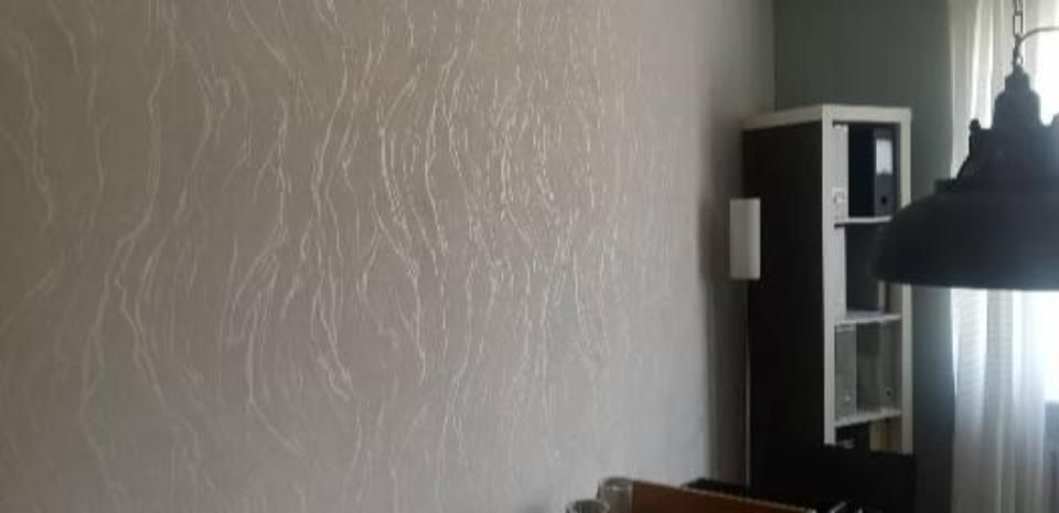 Graue, strukturierte Wand neben einem Fenster mit transparenten Vorhängen, schwarzer Hängelampe und einem weißen Regal mit Büchern.