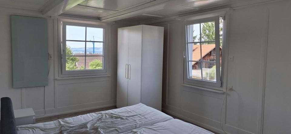 Kleines weisses Schlafzimmer mit zwei Fenstern, einem Kleiderschrank und einem ungemachten Bett.