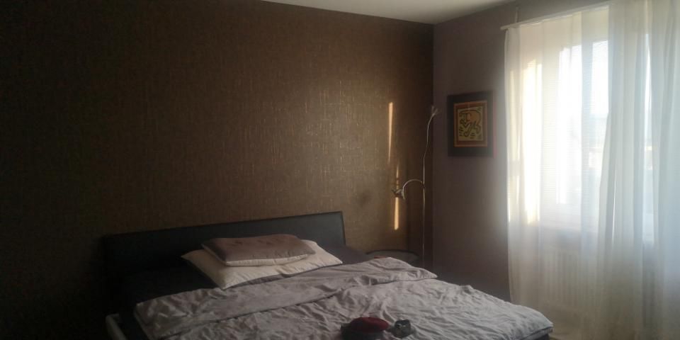 Dunkles Schlafzimmer mit ungemachtem Bett, braunen Wänden und Sonnenlicht durch dünne Vorhänge.