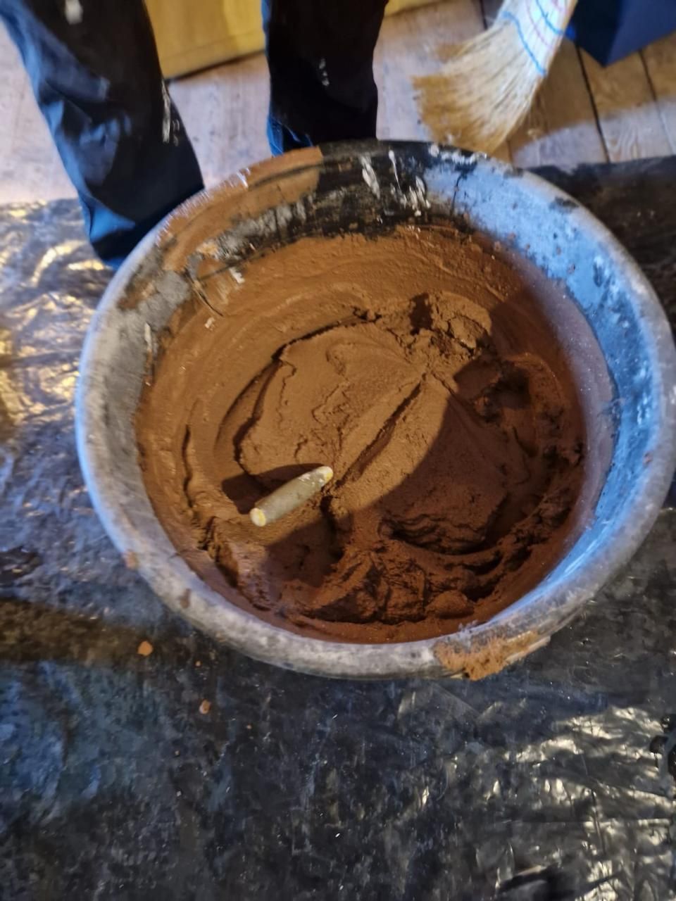 Eine Schüssel mit braunem Teig oder Lehm auf einem mit Plastikfolie bedeckten Boden, in der sich ein kleiner Holzstab befindet.