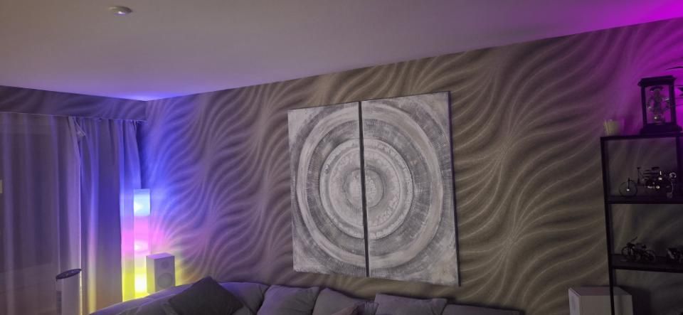 Wohnzimmer mit grauer, gewellter Wand, abstraktem Leinwandbild, violetten Akzentleuchten und einem schwarzen Regal rechts.