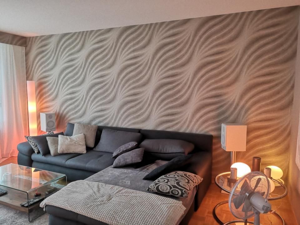 Modernes Wohnzimmer mit grauem Sofa, gemusterter Tapete, Glas-Couchtisch, Lampen und Standventilator