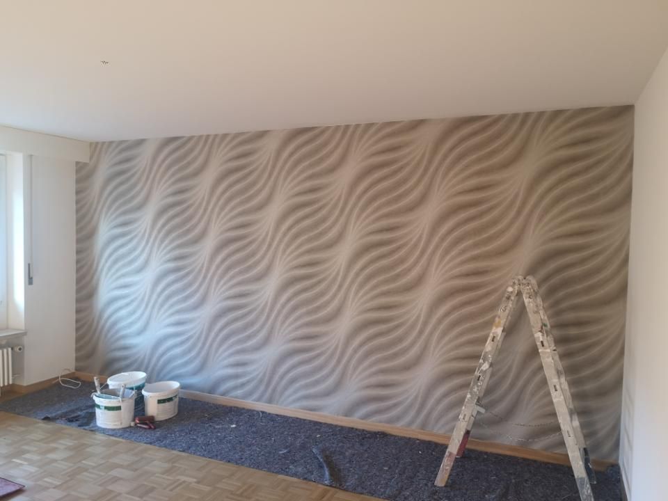 Eine teilweise gestrichene Wohnzimmerwand mit Wellenmustertapete, Farbdosen und eine Trittleiter auf dem Boden.