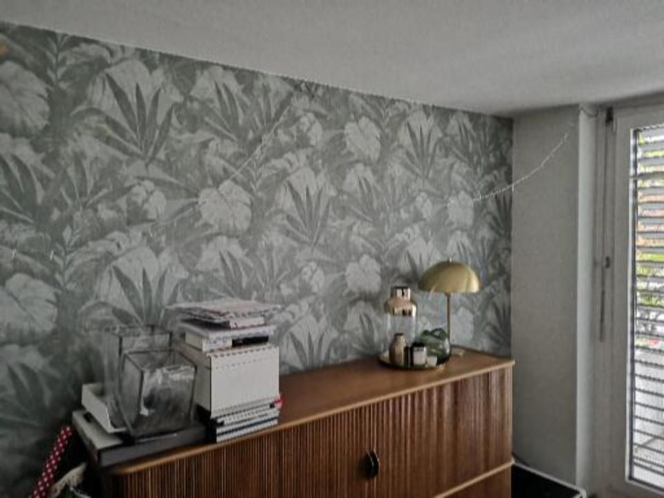 Kleines Zimmer mit tropischer Tapete, Holzkommode und einem Fenster mit Jalousien.