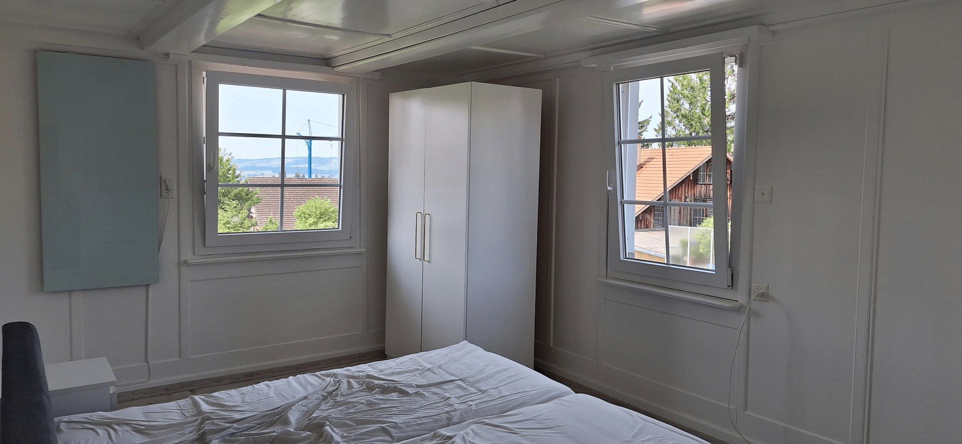 Helles Schlafzimmer mit weissem Kleiderschrank, Bett und zwei Fenstern mit Blick über die Dächer.