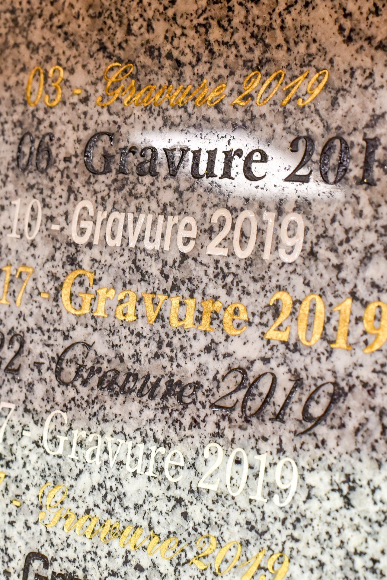 Gros plan sur un texte gravé sur une pierre mouchetée, avec la mention Gravure 2019 répétée en noir et or.