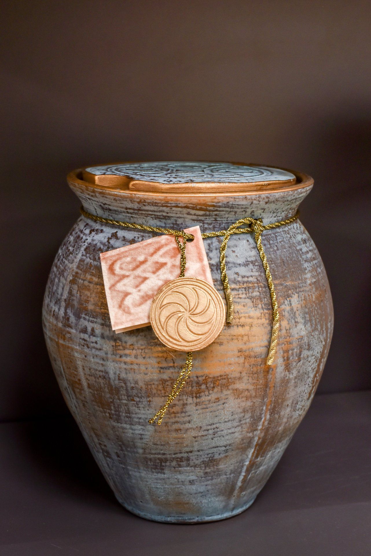 Vase rustique en céramique avec une chaîne et un pendentif en or, orné d'une note rose sur fond sombre.