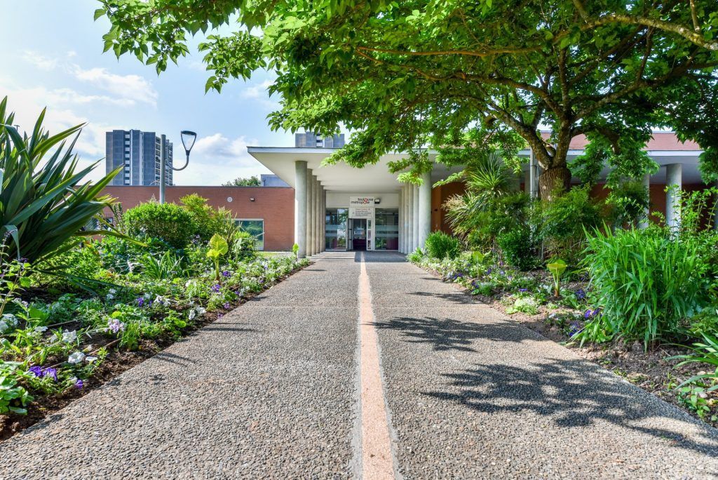 Allée paysagée menant à l'entrée d'un bâtiment moderne, bordée d'arbres et d'arbustes de part et d'autre.