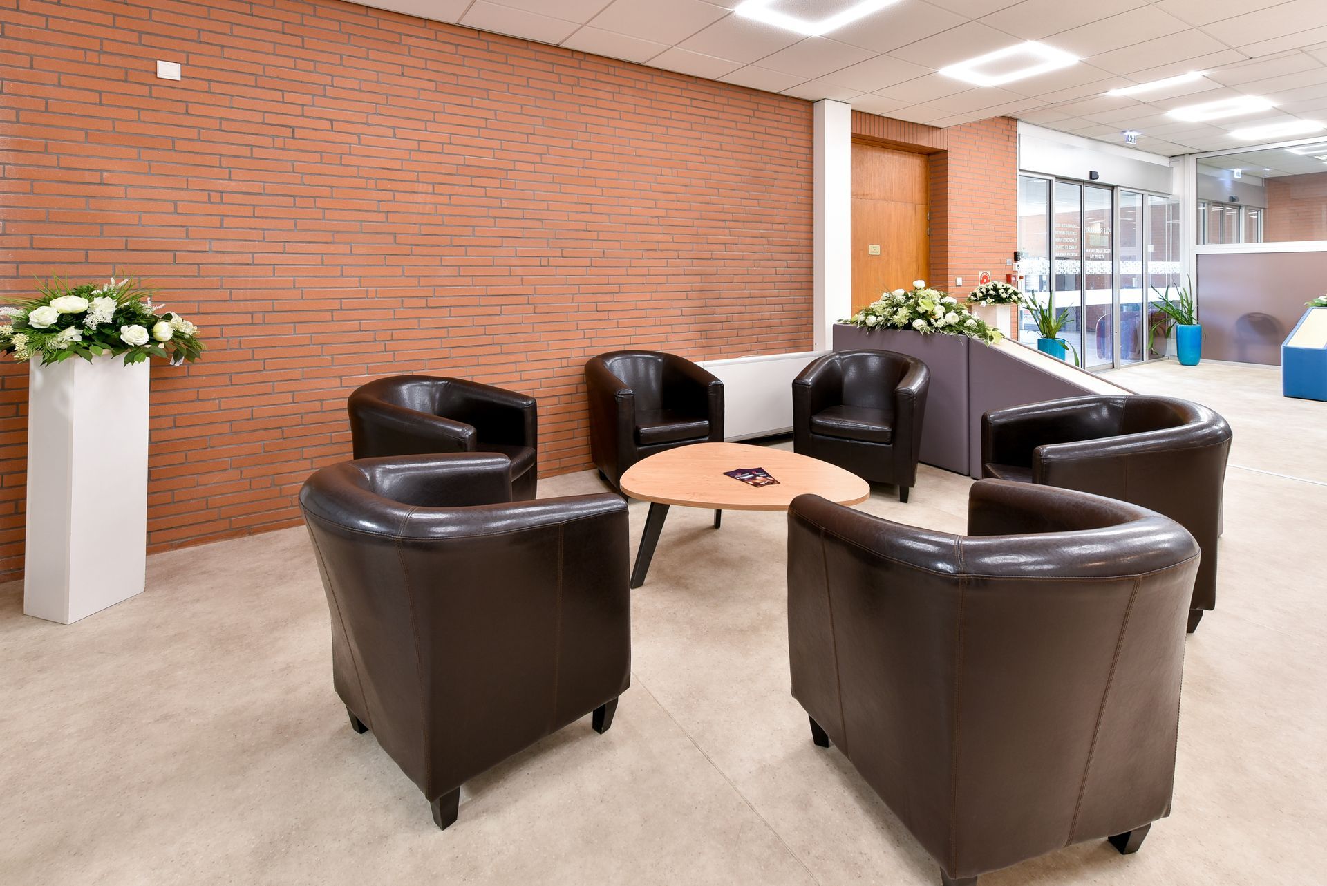 Un espace salon moderne dans le hall d'entrée, avec des fauteuils en cuir noir, des petites tables en bois et un mur de briques.