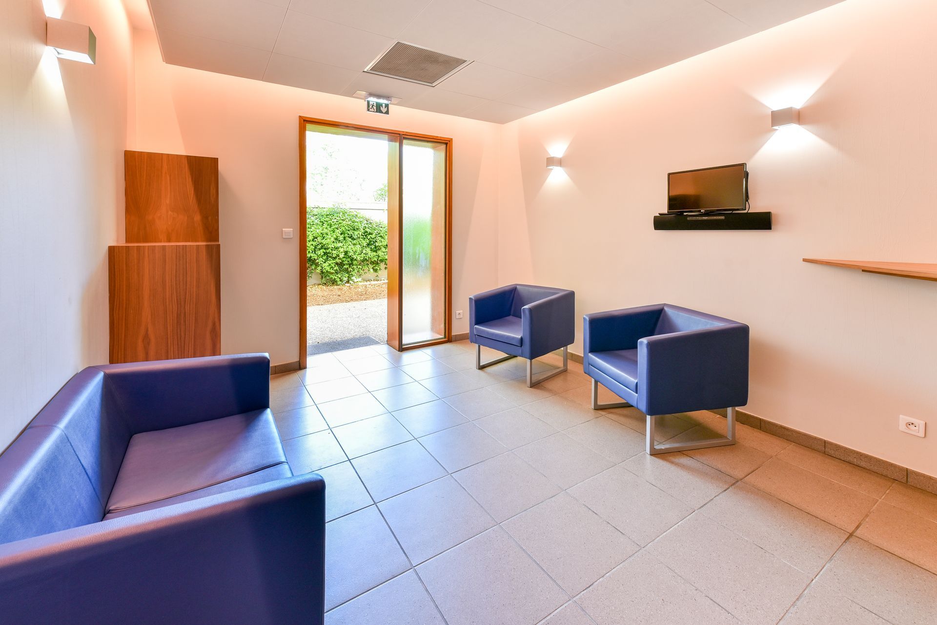 Salle d'attente lumineuse avec chaises bleues, sol carrelé, appliques murales et porte ouverte sur un espace vert.