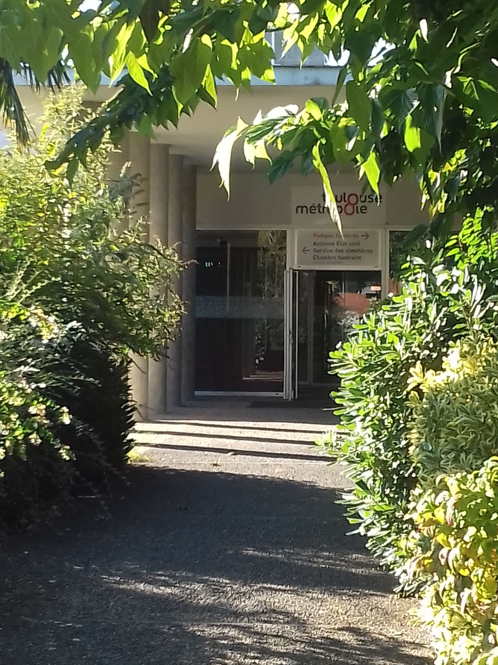 L'entrée du bâtiment, encadrée de verdure, donne sur des marches menant à une porte vitrée baignée de soleil.