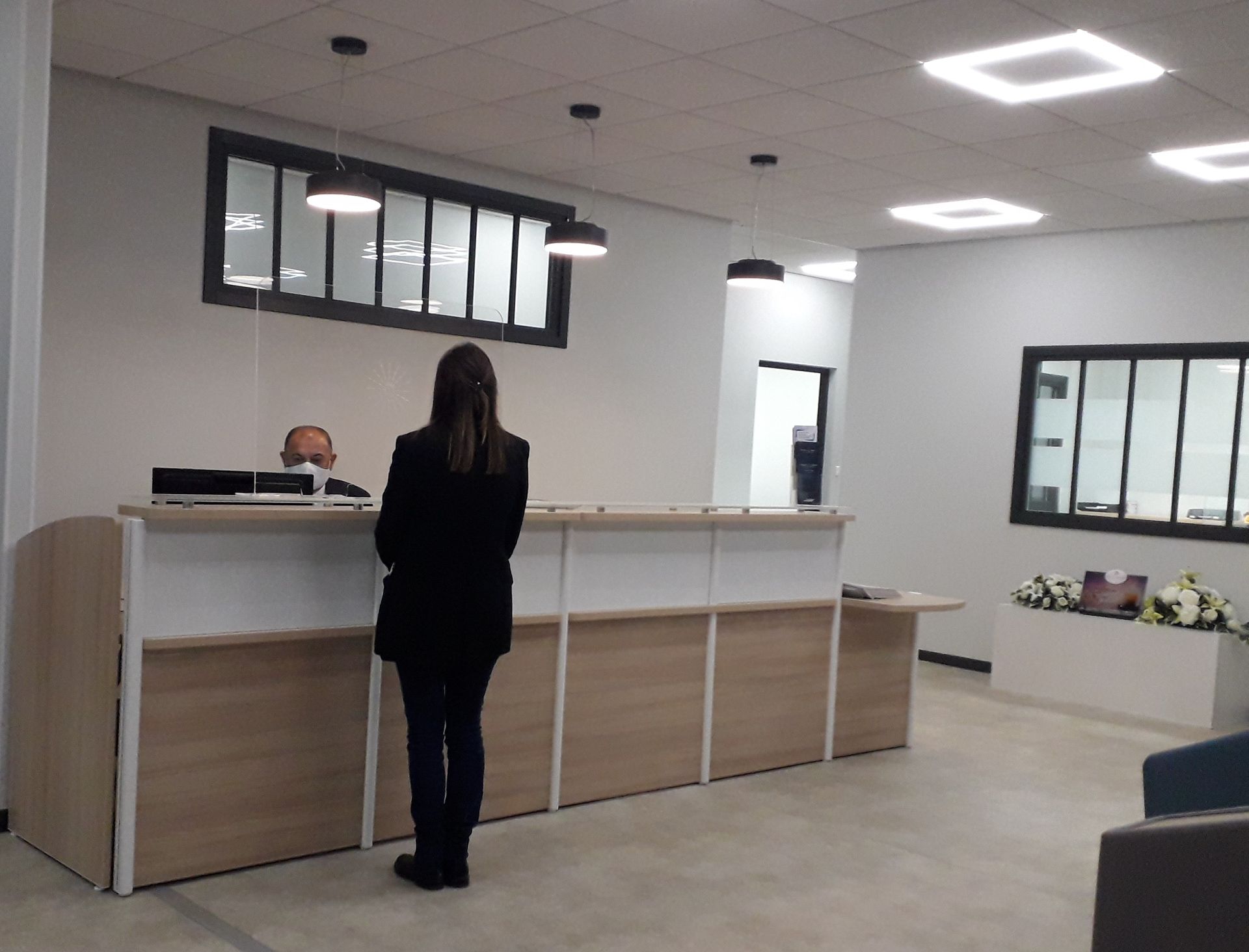 Une personne se tient debout derrière un comptoir d'accueil blanc dans le hall lumineux d'un immeuble de bureaux.