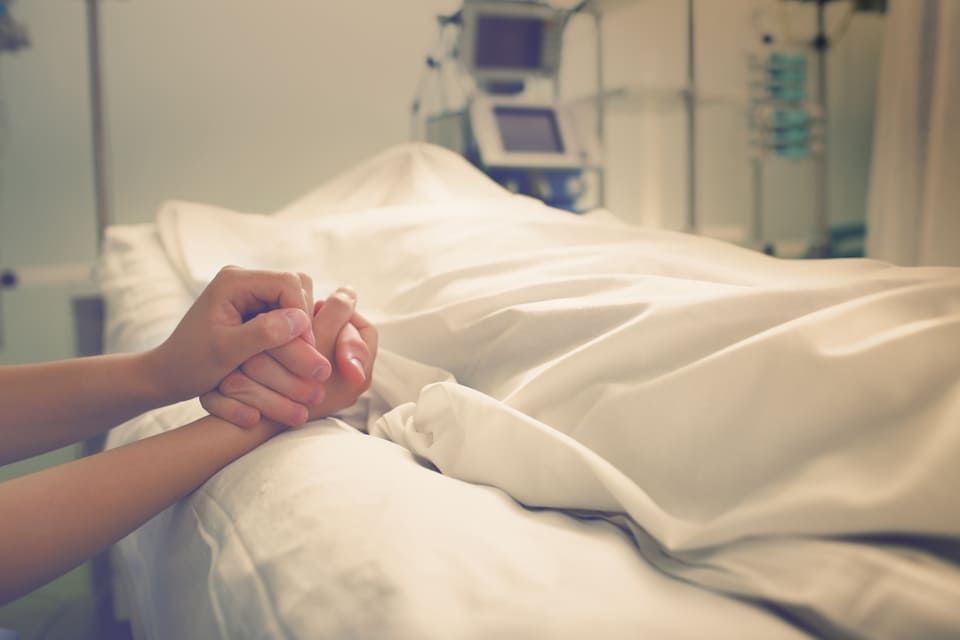 Mains jointes près d'un lit d'hôpital sous des draps blancs, suggérant réconfort ou soutien