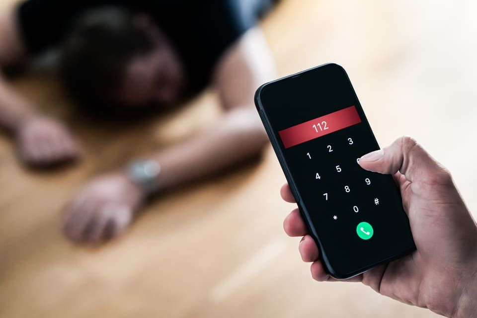 Main tenant un smartphone composant le numéro d'urgence 112, avec une personne blessée floutée en arrière-plan.