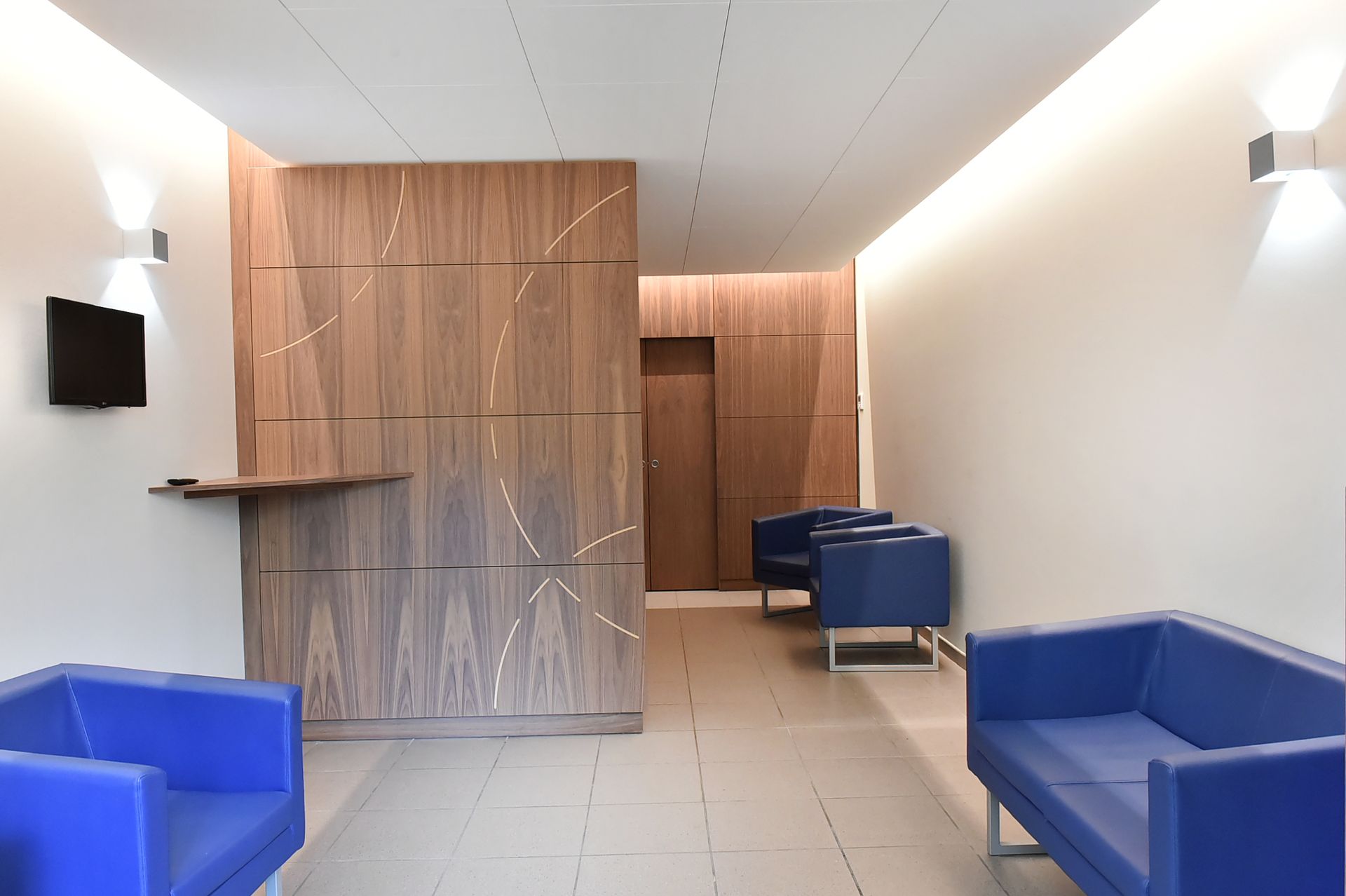 Couloir moderne avec chaises bleues, cloison en lambris et téléviseurs muraux.